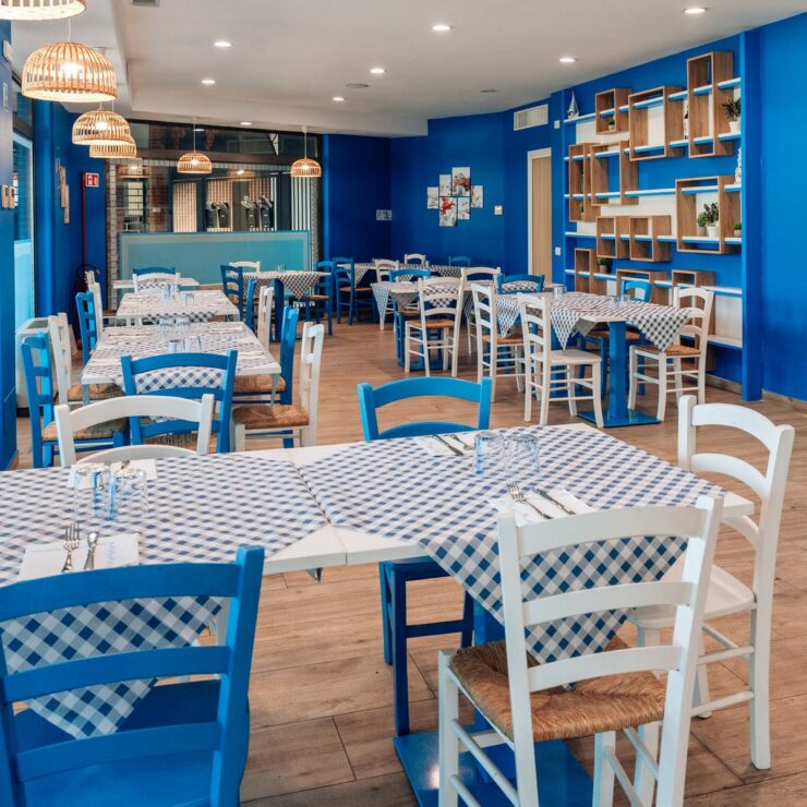 Sala di Olympos Cucina Greca a Limbiate con pareti blu, tovaglie a quadri e sedie impagliate