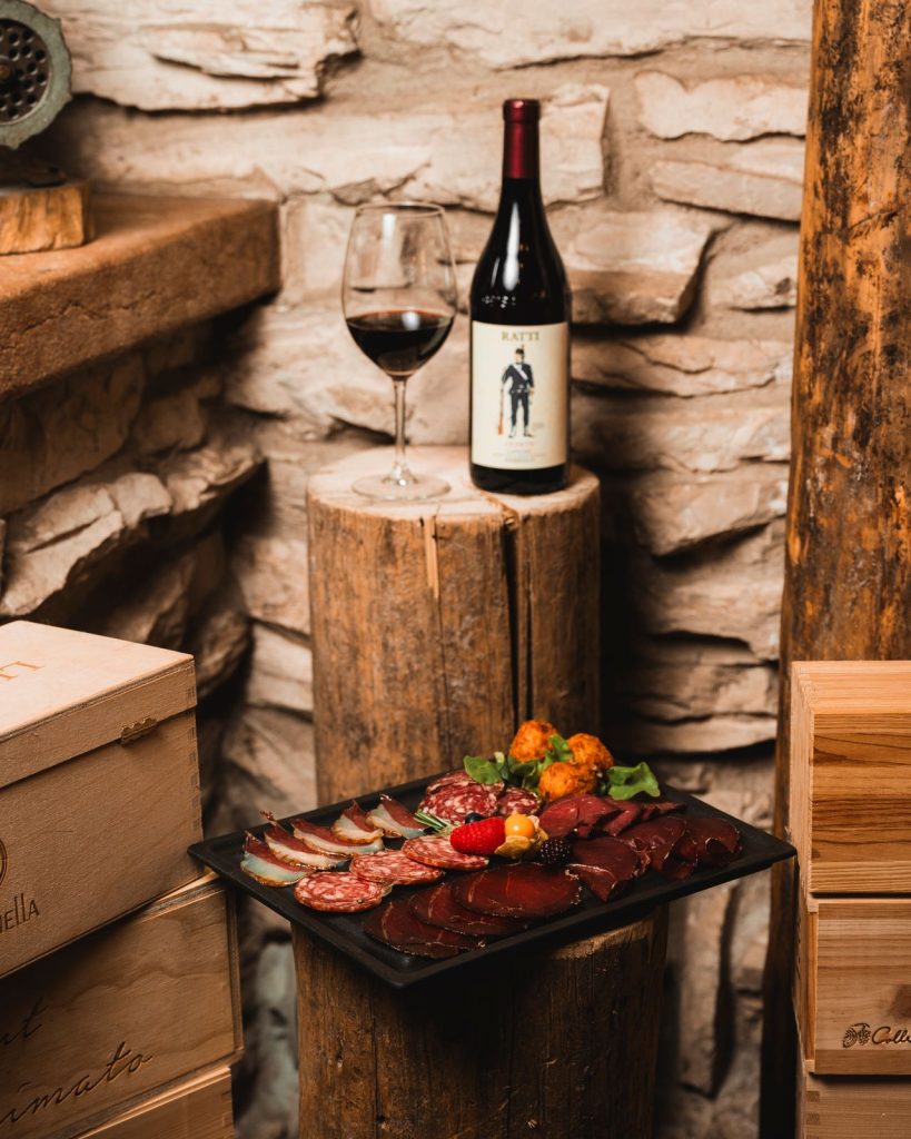 Bottiglia di vino rosso, calice e tagliere di salumi su un ceppo di legno davanti al muro in pietra